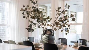 A dining area with a table set for two, featuring a vase with branches and leaves, and a cityscape view through large windows.