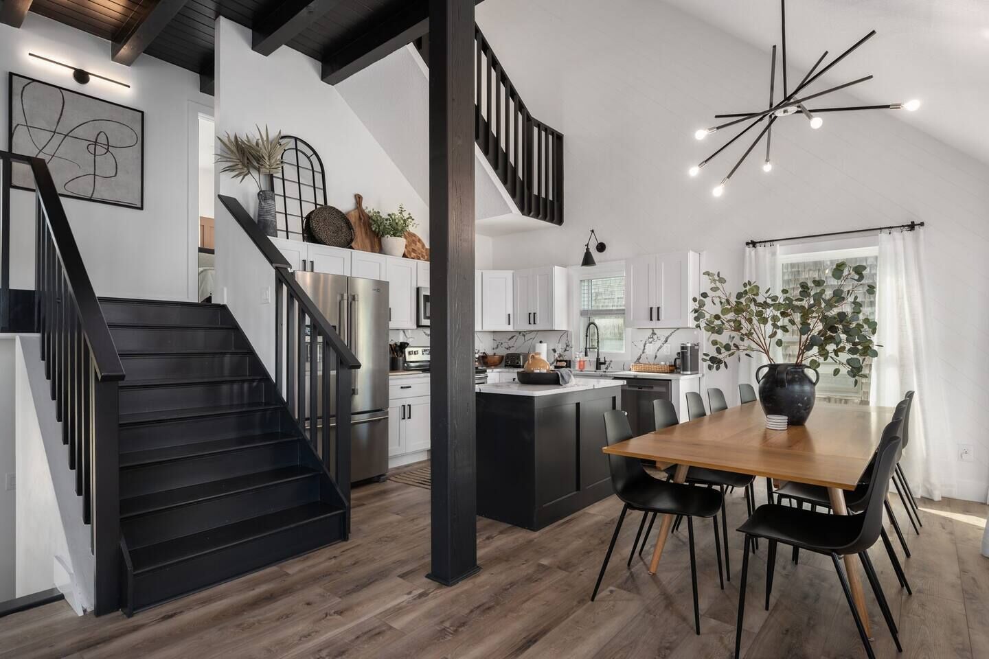 A modern kitchen with a central island, black staircase, and a dining area with a wooden table and black chairs.