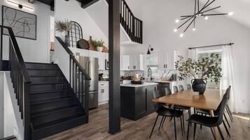 A modern kitchen with a central island, black staircase, and a dining area with a wooden table and black chairs.
