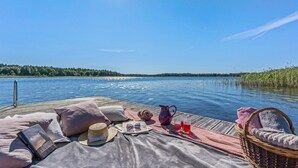 A wooden dock with a picnic setup including a blanket, pillows, a basket, a book, a hat, a pitcher, and glasses. - Awesome home in Köpmannebro with lake view (Köpmannebro)