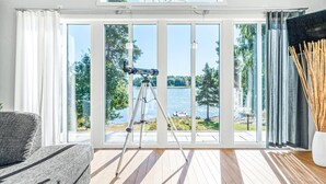 A room with large windows overlooking a lake, a telescope on a tripod, and a view of trees and a dock. - Awesome home in Köpmannebro with lake view (Köpmannebro)