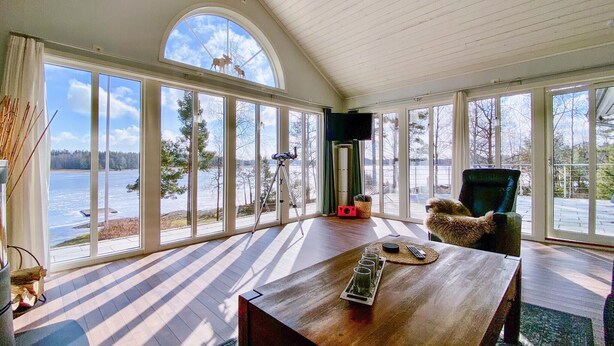 A spacious room with large windows overlooking a lake, a wooden table with glasses and a remote, and a telescope on a tripod. - Awesome home in Köpmannebro with lake view (Köpmannebro)