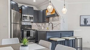 Une cuisine moderne avec des meubles de couleur sombre, des appareils en acier inoxydable et un pare-feu en marbre. On y trouve également un coin repas avec une table, des chaises et une plante en pot.
