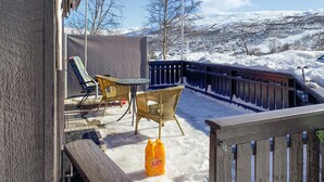A covered outdoor seating area with wicker chairs and a table, overlooking a snowy landscape with mountains in the background.