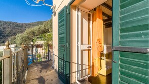 Un étroit couloir extérieur mène à une pièce dotée de volets verts, d’une porte et d’un petit balcon offrant une vue sur les collines.
