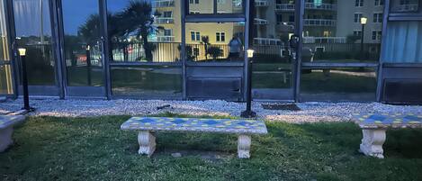 Un patio con dos bancos de piedra y una mesa con azulejos, rodeado por un muro de piedra bajo y un área con césped.