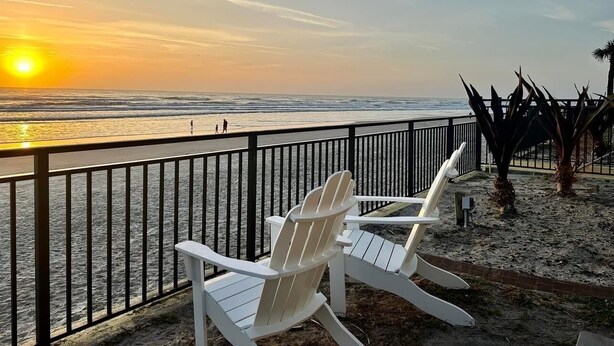 A beach view with two white Adirondack chairs facing the ocean, a sandy walkway, and a palm tree. - Friends & Family Beach Escape - Double Oceanview Studio Sleeps up to 10 (Daytona Beach)