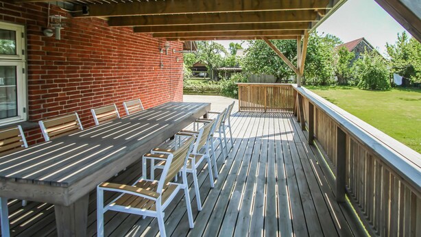 Une terrasse couverte avec une table et des chaises en bois, un mur en briques et une vue sur une pelouse verdoyante et des arbres.