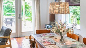 Een eethoek met een tafel gedekt voor twee personen, een rieten lamp en een vaas met bloemen.