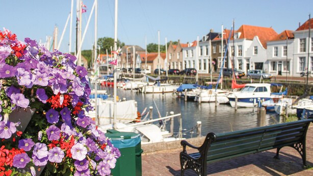 Een jachthaven met aangemeerde boten, een groene bank en een bloembed met paarse en rode bloemen.