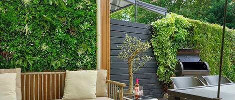 A patio with a wooden bench, a small table with a bottle and glasses, and a grill.