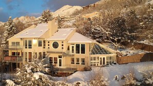 Una casa moderna de dos pisos con un balcón grande, rodeada de árboles nevados y montañas al fondo.