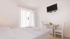 Quarto com cama, escrivaninha, cadeira e televisão fixada na parede.