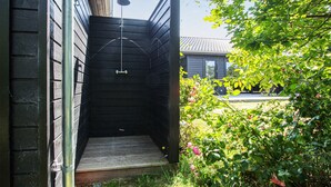 A wooden outdoor shower area with a metal showerhead, a small wooden step, and a garden with green foliage and pink flowers. - Awesome home in Samsø with WiFi (Samsø)