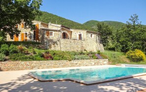 Une maison en pierre avec une piscine, entourée d’une végétation luxuriante et de collines.