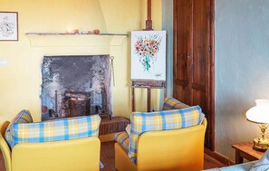 Ein Zimmer mit Kamin, zwei sesseln mit Karomuster, einem Bild an der Wand und einer Holztür.