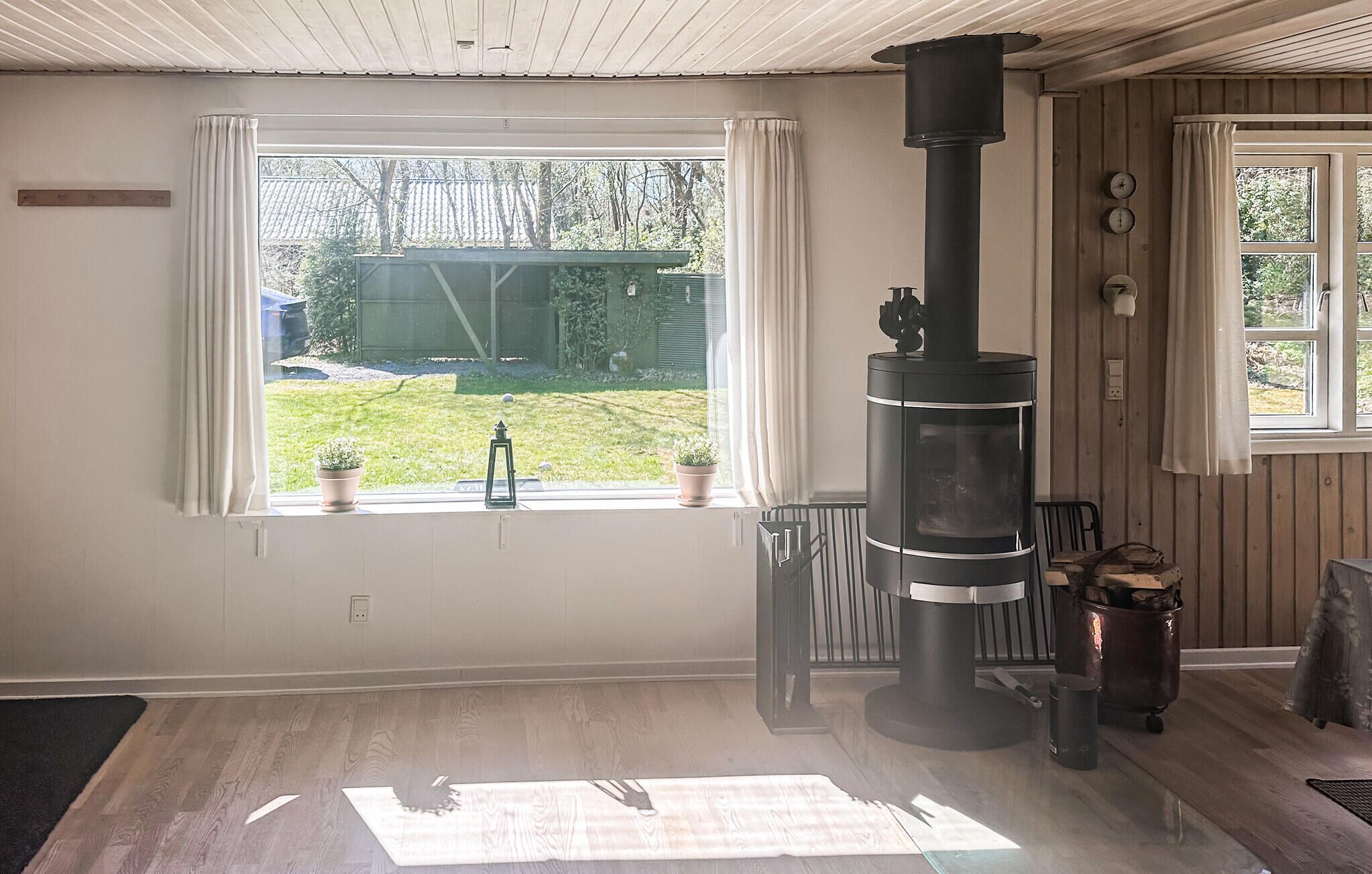 Ein Zimmer mit einem großen Fenster, einem Kaminofen, einem Blumentopf und Blick auf einen Garten.