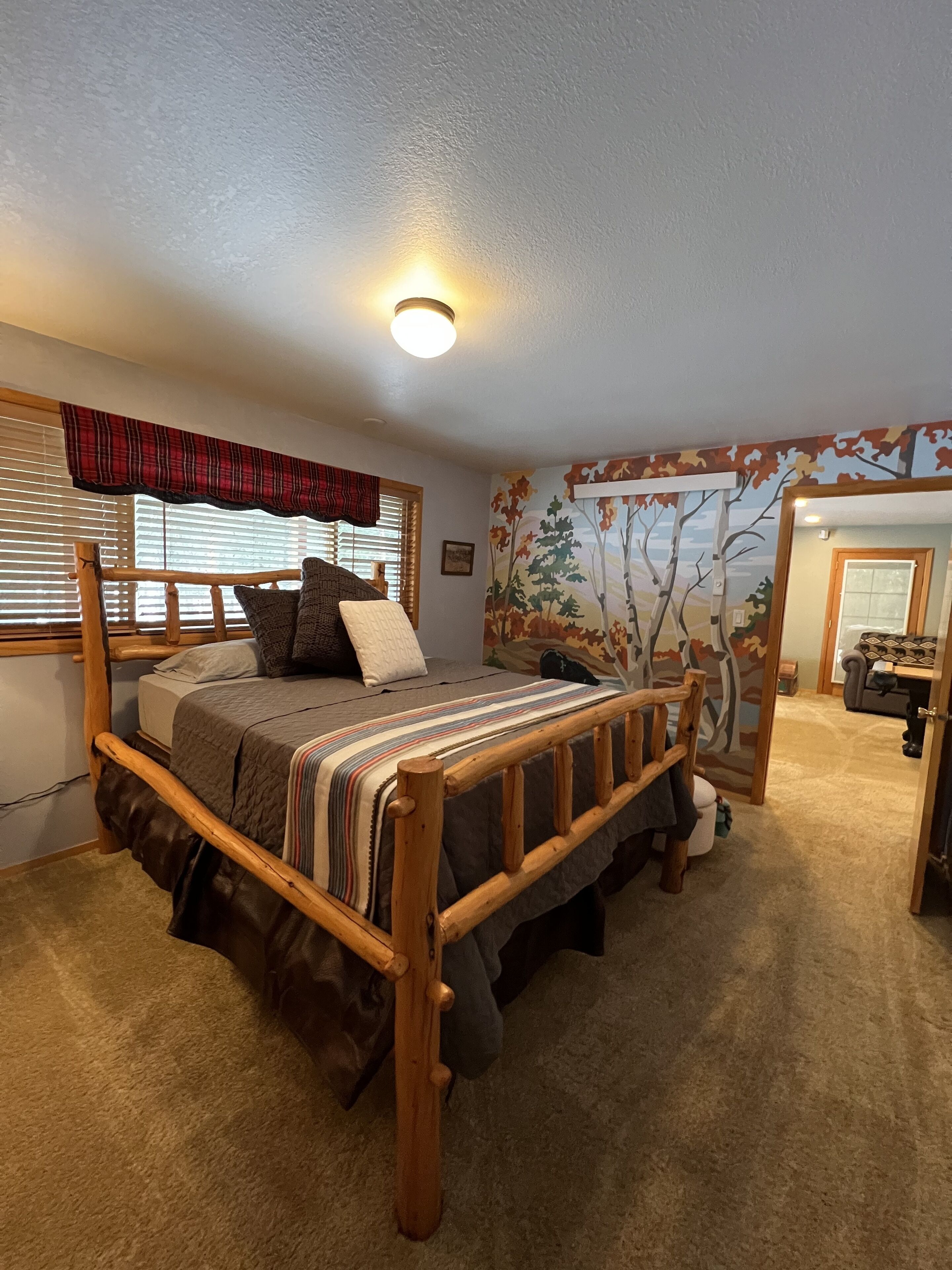 A bedroom with a wooden bed, a window with blinds, a wall mural, and a carpeted floor.