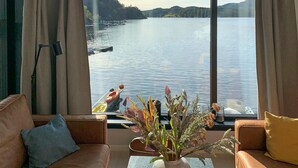 A room with a large window overlooking a lake, a sofa, a coffee table, and a vase with dried plants.