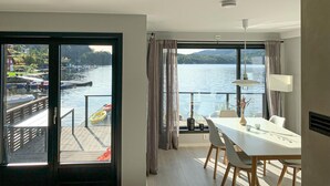 A modern room with a large window overlooking a lake, a dining area with a table and chairs, and a view of boats and docks outside.