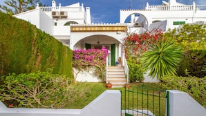 A white building with a staircase leading to a door, surrounded by greenery and flowering plants.