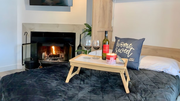 Une chambre avec une cheminée, un lit recouvert d’une couverture sombre, une table de chevet avec une bougie, une bouteille de vin et une carafe.