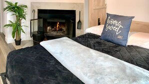 Une chambre avec une cheminée, un lit recouvert d’une couverture sombre, un oreiller bleu orné du texte « chez soi, quel bonheur », et une plante en pot.