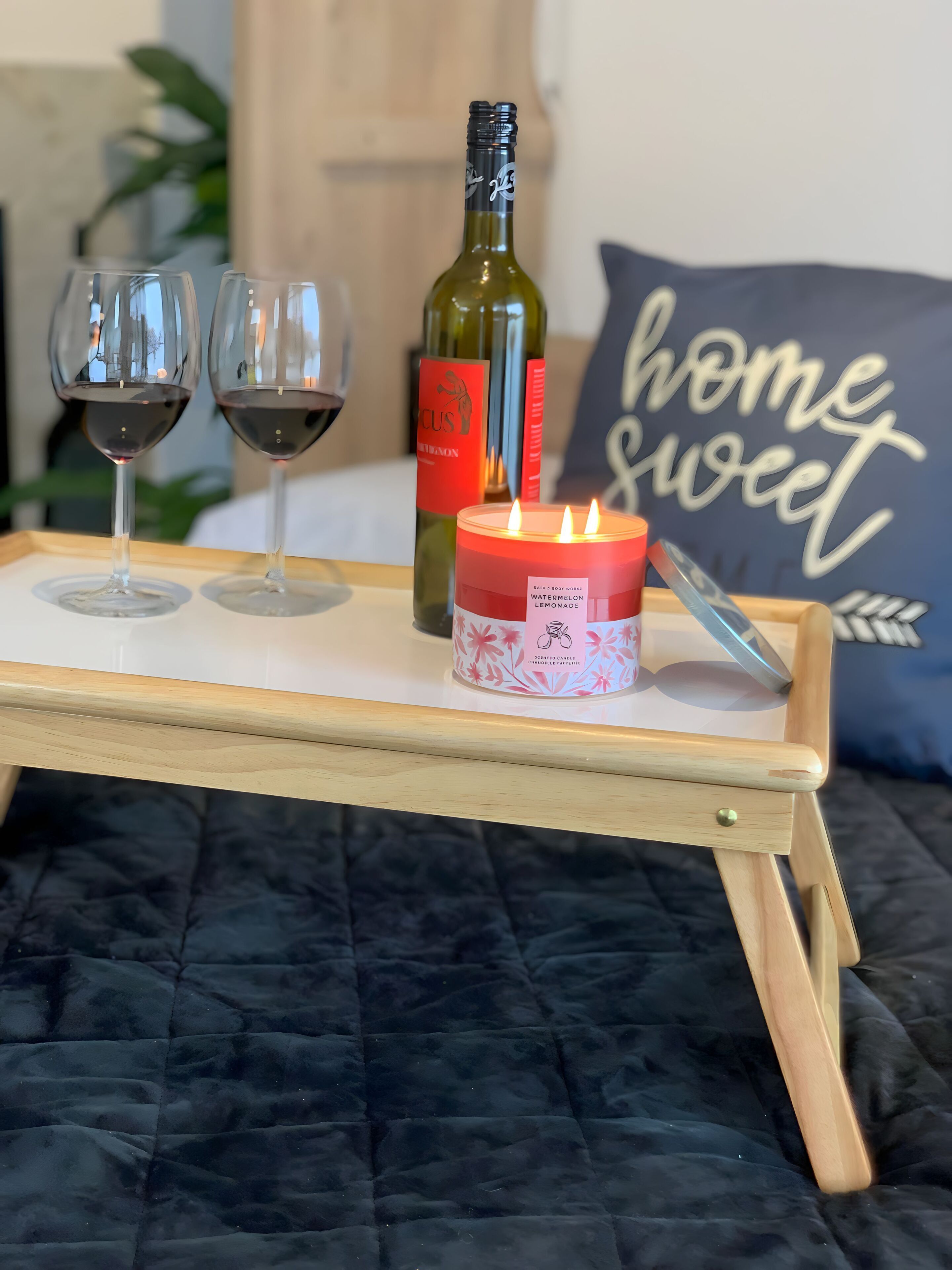 Un plateau avec deux verres à vin, une bougie et une bouteille de vin, posé sur un lit recouvert d’une couverture sombre.
