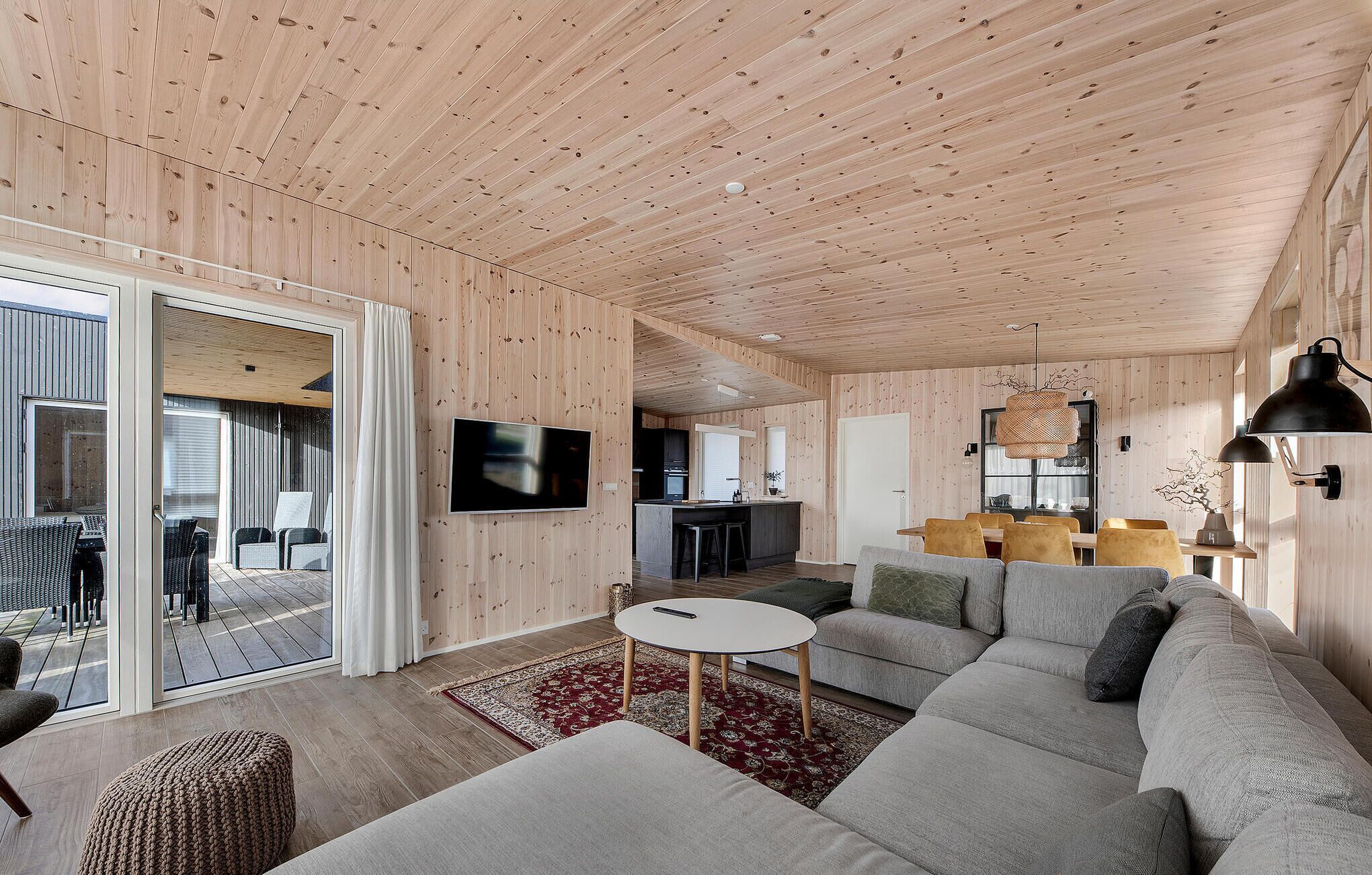 A modern interior with a wooden ceiling, a large sofa, a dining area, and a kitchen in the background.