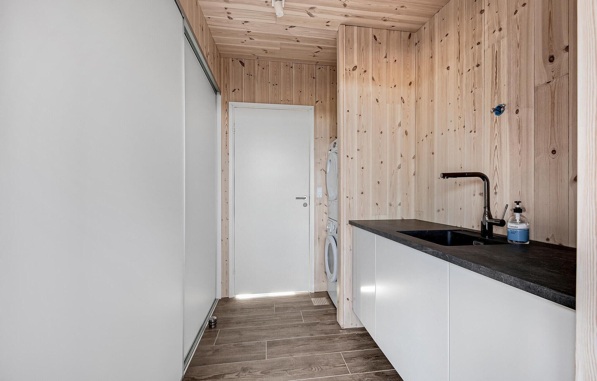 A narrow hallway with a wooden ceiling and walls, leading to a white door. A black countertop with a sink and a bottle of hand sanitizer is visible.