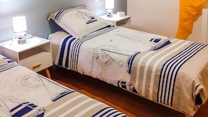 Habitación de hotel con dos camas, cada una con cubrecama y almohadas con motivos náuticos. Hay mesitas de noche con lámparas y un pequeño cajón.