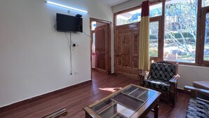 Ein Wohnzimmer mit an der Wand montiertem Fernseher, einem Glastisch und einem gemusterten Sessel, mit Blick ins Freie durch große Fenster.