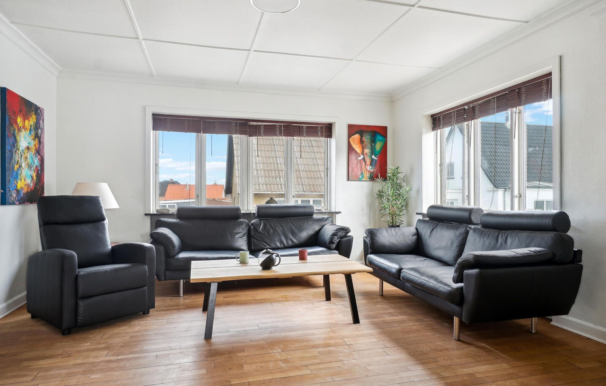 A living room with a sofa set, a coffee table, and two armchairs. There are two windows with curtains, a painting on the wall, and a potted plant.