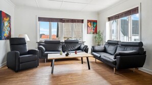 Ein Wohnzimmer mit einer Sofagarnitur, einem Couchtisch und zwei Sesseln. Zwei Fenster mit Vorhängen, ein Bild an der Wand und ein Blumentopf sind zu sehen.