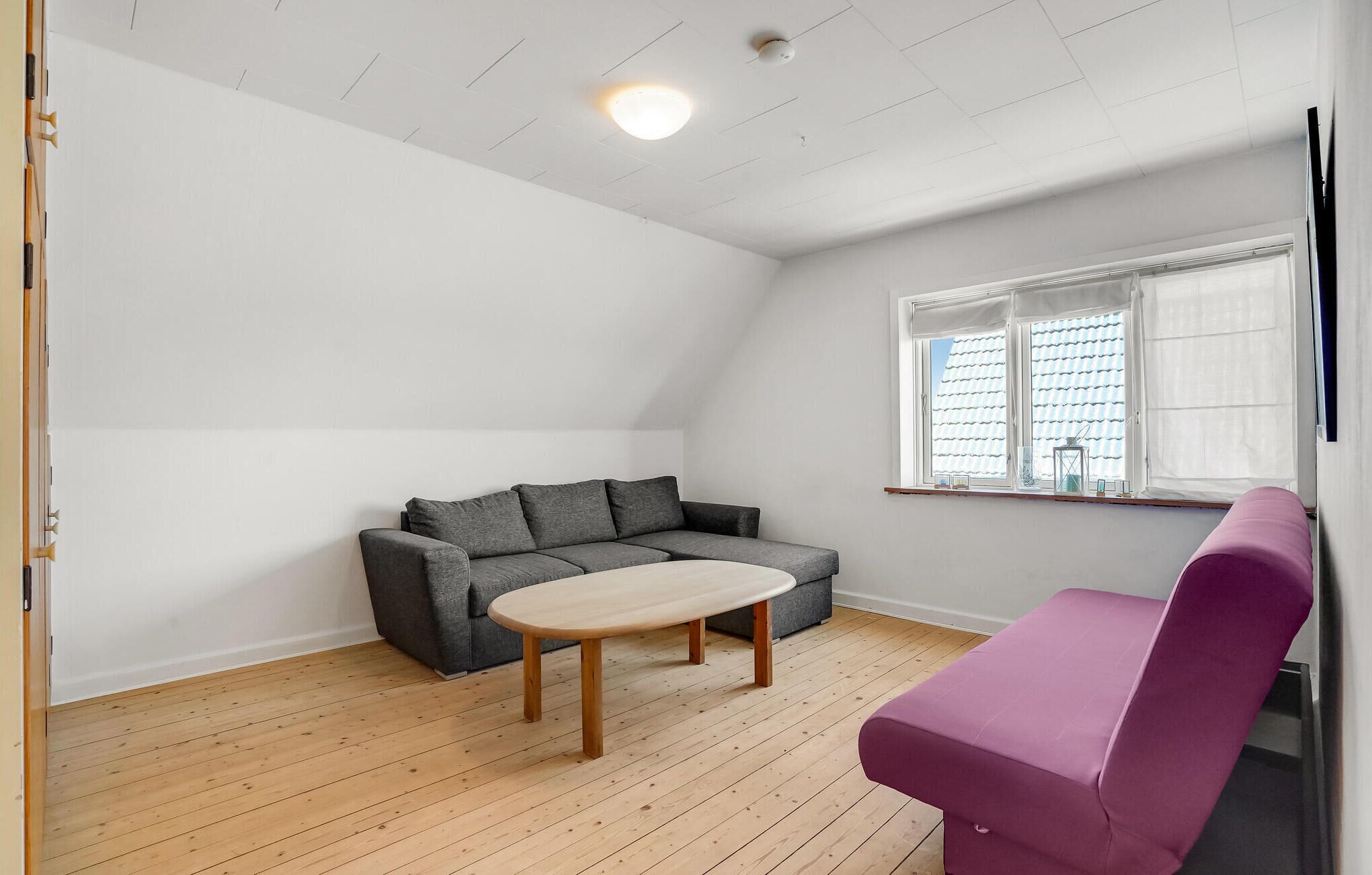 A room with a grey sofa, a small round table, a purple bench, and a window with white blinds.
