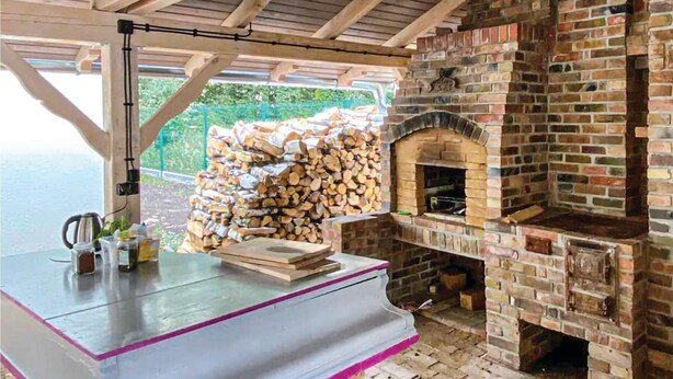 Une cuisine d’extérieur rustique, avec un four en briques, une pile de bois de chauffage et une table sur laquelle se trouvent une bouilloire et des salières.