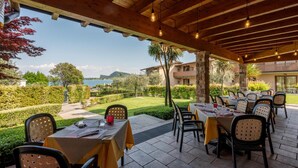 An outdoor dining area with tables and chairs, overlooking a landscaped garden and a body of water. - Beach front apartment (Manerba del Garda (BS))