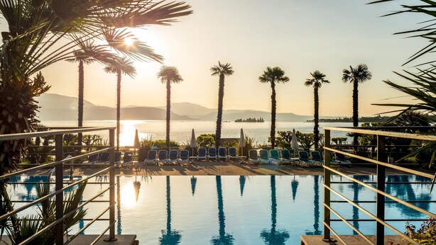 A pool area with lounge chairs and palm trees, overlooking a body of water and distant mountains. - Beach front apartment (Manerba del Garda (BS))