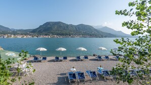 A beach with rows of blue lounge chairs and white umbrellas, a pebble beach, and a view of a lake with mountains in the background. - Beach front apartment (Manerba del Garda (BS))