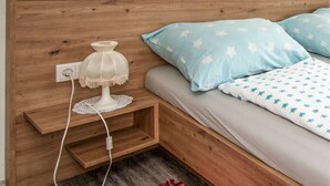 A wooden bed with a light blue blanket and pillows, a bedside table with a lamp, and a power outlet.