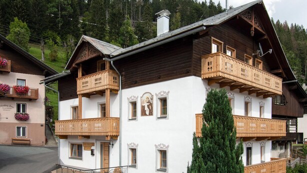 A traditional wooden chalet with balconies, a central staircase, and a mountain backdrop.