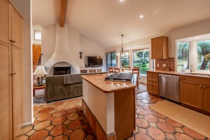 A modern kitchen with a stone floor, wooden cabinets, a built-in stove, and a large window overlooking a garden.