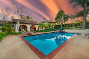 A swimming pool with a red brick border, surrounded by palm trees and a house with arched windows.