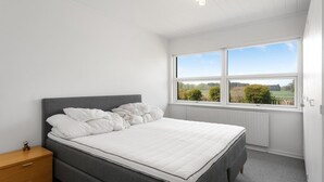 Ein Schlafzimmer mit Bett, Nachttisch, Fenster und Blick auf Grünflächen sowie einer Deckenleuchte.
