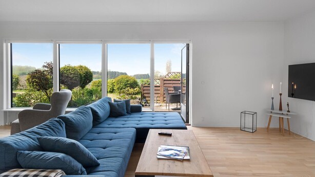 Ein modernes Wohnzimmer mit einem großen, blauen Ecksofa, einem Holztisch und Blick auf einen Garten durch große Fenster.
