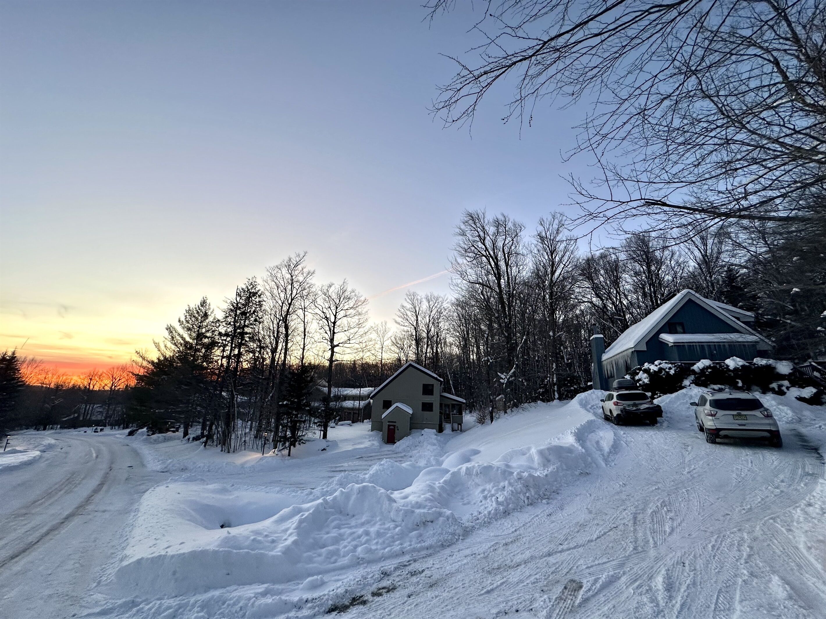 A snow-covered road with a house and a small cabin in the background.