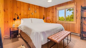 Une chambre avec des murs en bois, un lit recouvert de draps blancs, un banc, une table de chevet, une lampe et une fenêtre donnant sur un paysage verdoyant.