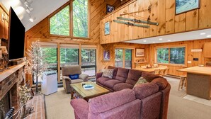 L’intérieur chaleureux d’un chalet avec une cheminée, des sièges confortables et de grandes fenêtres donnant sur la forêt environnante.