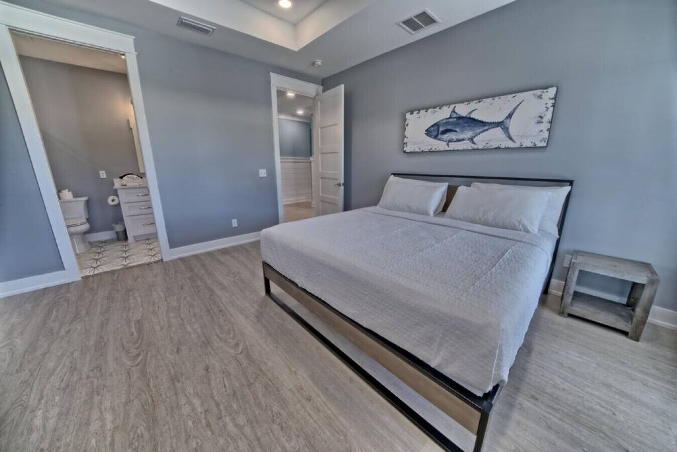A bedroom with a bed, a nightstand, a small table, a painting of a fish, and a bathroom visible through an open door.