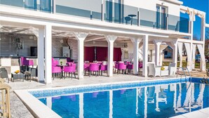A poolside area with a clear blue pool, white columns, and pink chairs.